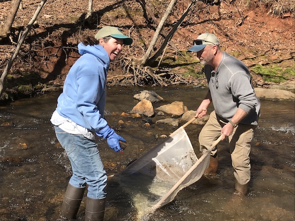 two volunteers in a stream, smiling and using a kick net to sample aquatic life