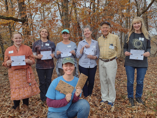 six people posed outdoors with paper certificates and one person kneeling in front of them holding nature journals