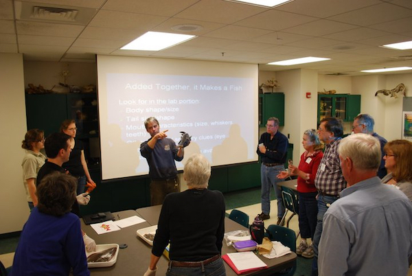 teacher in front of a screen, pointing out features of a fish to a classroom of a dozen adults