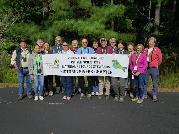 fifteen people standing outdoors in front of a tree, holding a banner reading "Volunteer Educators, Citizen Scientists, Natural Resource Stewards, Historic Rivers Chapter" with the Virginia Master Naturalist logo