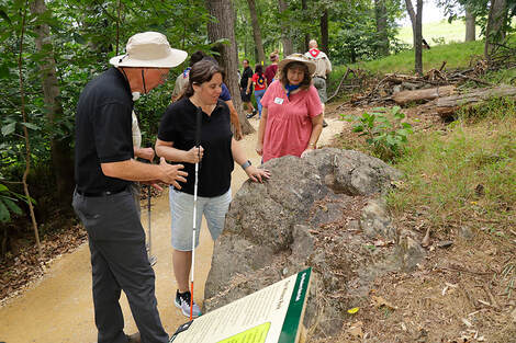 Woman holding white cane, touching a large boulder alongside a walking trail in a forest, while two volunteers speak to her about geology.