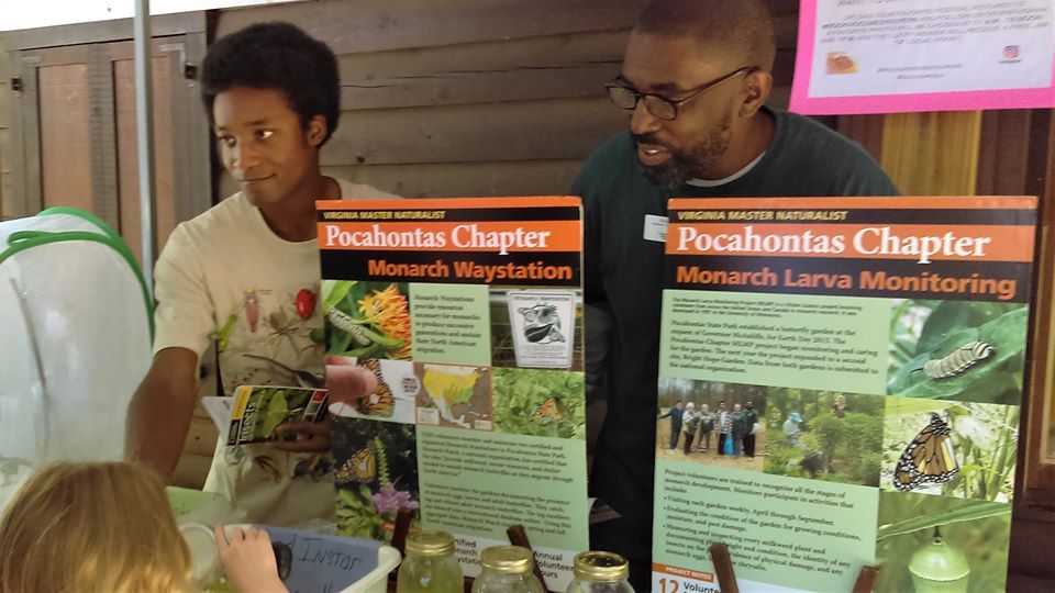 Two people at a display table with information about monarch butterflies, talking to a child looking through a magnifying glass