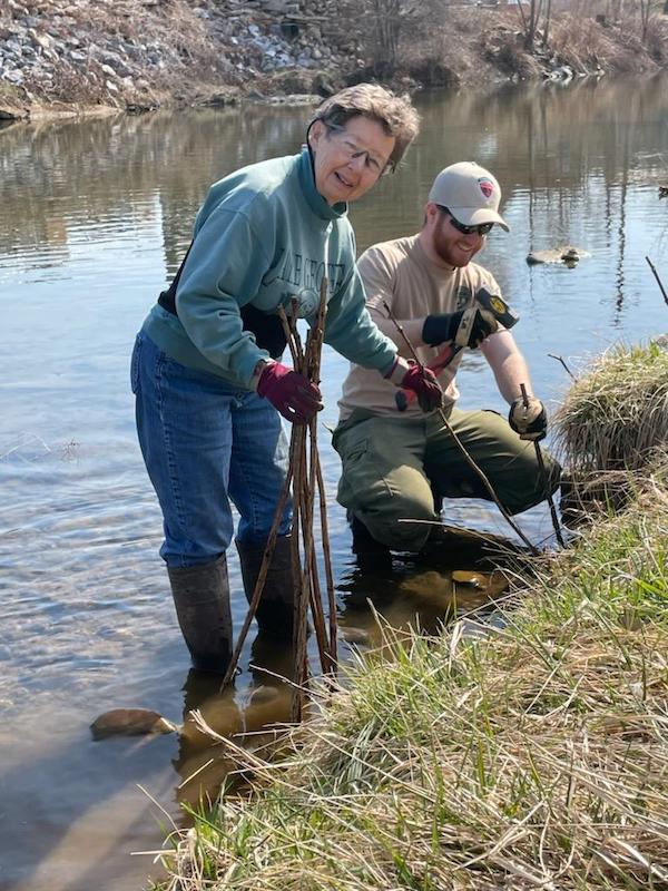 volunteer works alongside a forester to plant trees along a stream bank