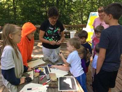 Adult instructing 9 children and one adult by a table covered in paper, pens, drawings, and maps.