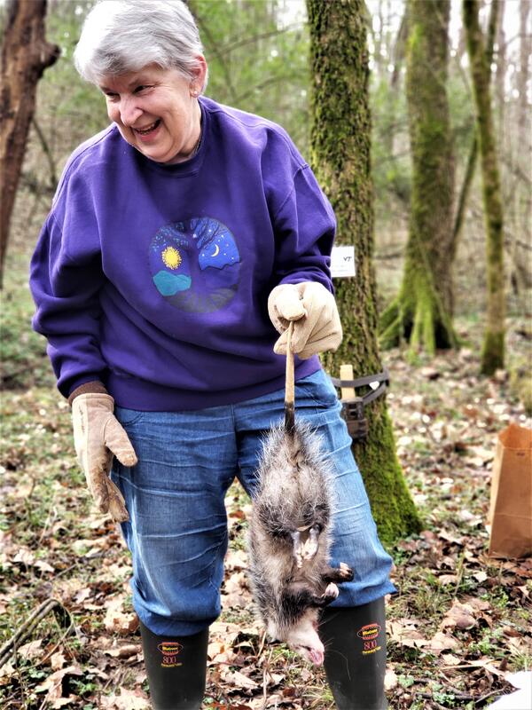 A smiling woman in a forest, wearing boots and gloves, holding a dead opossum by the tail