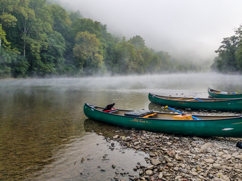 Three green canoes landed on rocky river shoreline with mist rising over the river and green forested hill in background