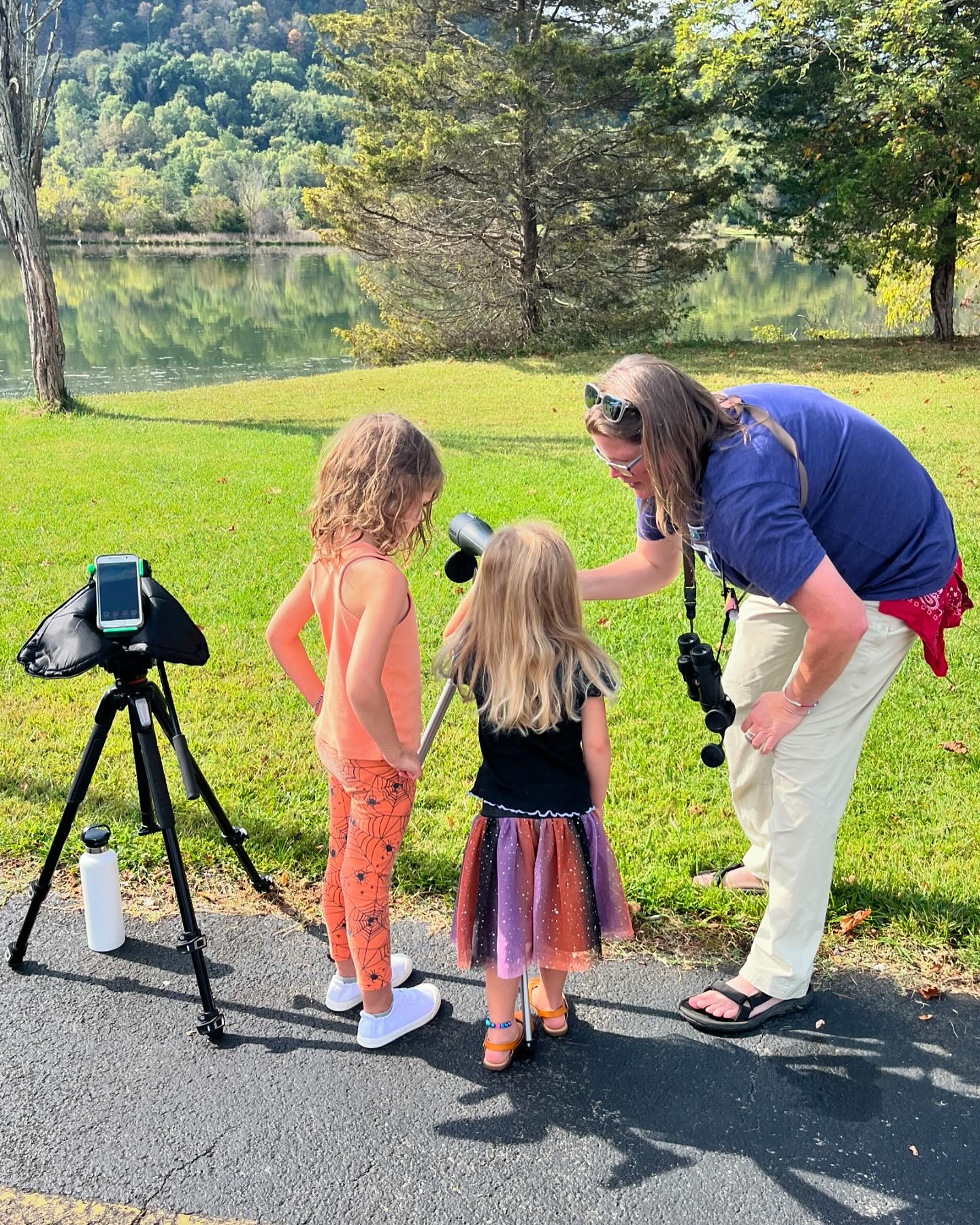 adult volunteer next to a lake, showing two young children how to look through a spotting scope